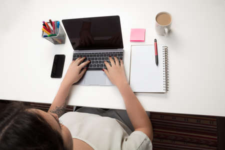 High angle view of a young woman using a laptop computer seated at a table with a notebook, mobile phone and coffeeの写真素材