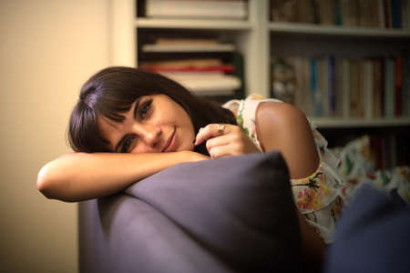 Thoughtful attractive young woman gazing at camera as she leans on the back of a comfortable sofa resting her head on her armの写真素材