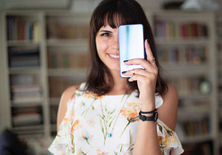 Young smiling cheerful brunette woman looking to camera while holding brand new smartphone in hand...