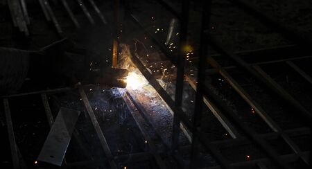 A welder at work in a workshop produces metal structures. Sparks from welding fly around.