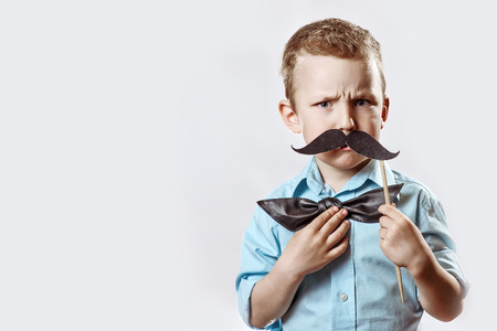 a frowning serious boy in a light shirt put a mustache on a stick and a bow tie on his face to make him look older.の写真素材