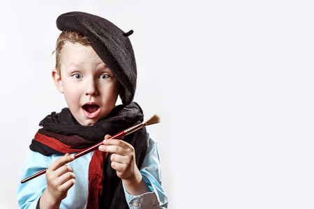 cheerful boy artist in black beret, scarf and with a brush in his mouth on a light backgroundの写真素材