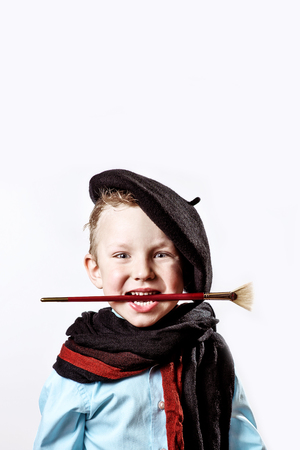cheerful boy artist in black beret, scarf and with a brush in his mouth on a light backgroundの写真素材