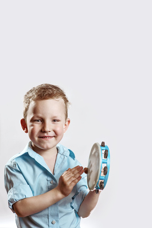 cheerful boy in a blue shirt holding a tambourine and smiling on a light backgroundの写真素材