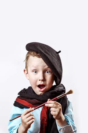 cheerful boy artist in black beret, scarf and with a brush in his mouth on a light backgroundの写真素材