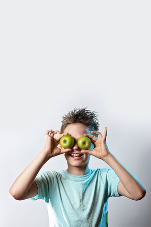 a boy in a blue t-shirt put two green apples to his eyes and laughs on a light backgroundの写真素材