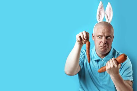 Bald brutal man in a light t-shirt and rabbit ears. In his hands he holds the carrot of a different...