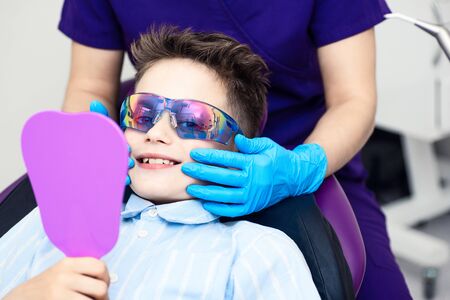 the boy in special glasses in the dentist's chair. the doctor in sterile gloves shows him his smile in the mirror.の写真素材