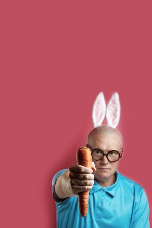 Bald brutal man in a light t-shirt and rabbit ears. In his hands he holds the carrot like a dumbbell