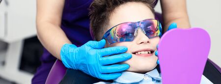 the boy in special glasses in the dentist's chair. the doctor in sterile gloves shows him his smile in the mirror.の写真素材