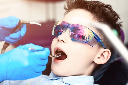 the boy in special glasses in the dentist's chair. the doctor in sterile gloves shows him his smile in the mirror.の写真素材