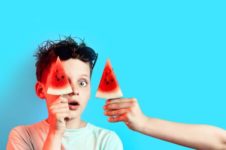 boy in a light t-shirt watermelon on a stick closes one eye on a blue backgroundの写真素材