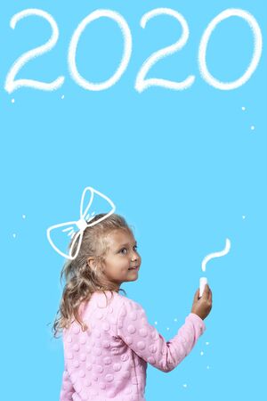 Cute girl with white chalk on blue background. Writes on the Board 2020 yearの写真素材