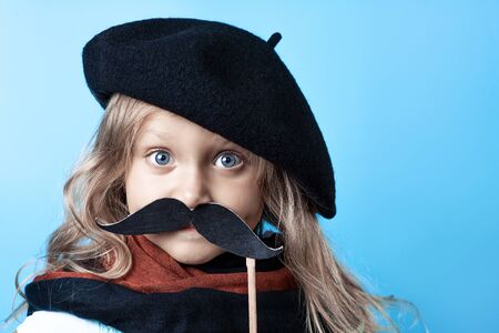 Funny girl in black beret, scarf and mustache on a stick on a blue backgroundの写真素材