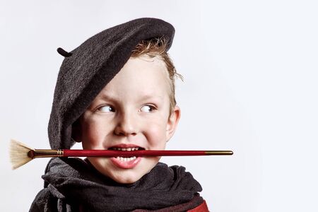 cheerful boy artist in black beret, scarf and with a brush in his mouth on a light backgroundの写真素材