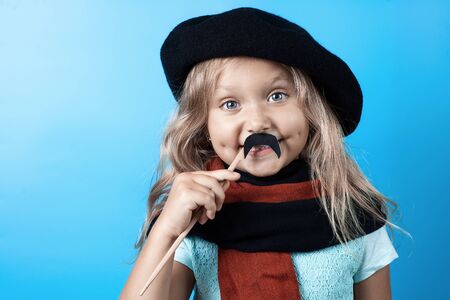 Funny girl in black beret, scarf and mustache on a stick on a blue backgroundの写真素材