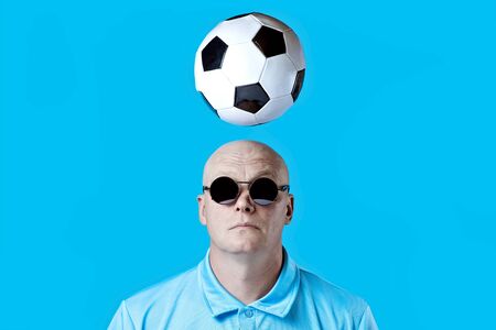 bald brutal man in dark round glasses with highlights. A football hovered overhead. Blue background.の写真素材