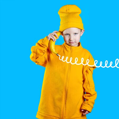 A cool boy in a bright yellow jacket and hat holds a banana in his hands like a phone. On blue...