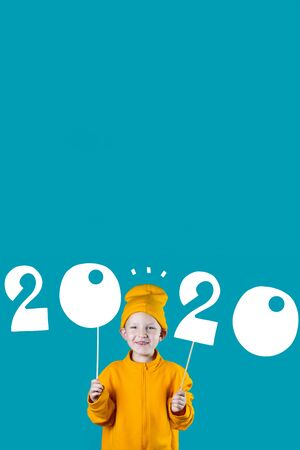 cheerful boy in a yellow hat and jacket holding the figures 2020 on bright colored background