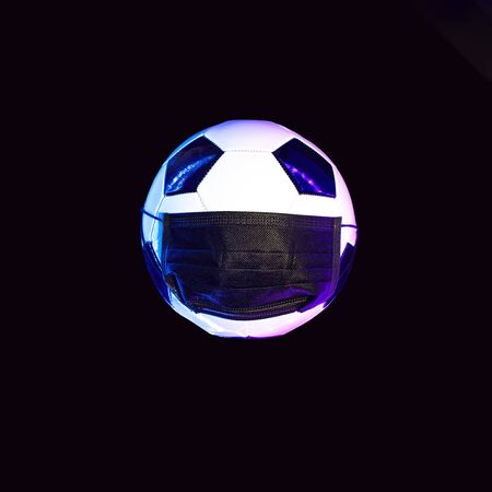 A soccer ball in a black medical mask from the virus. In the light of neon on a dark background.