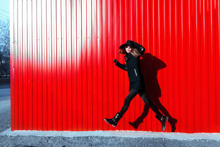 Running girl in a black hat, glasses, coat against a bright red wall.の写真素材