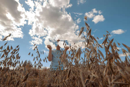 A boy with two round mirrors, in which the sky is reflected in glasses, stands in a field of wheat.