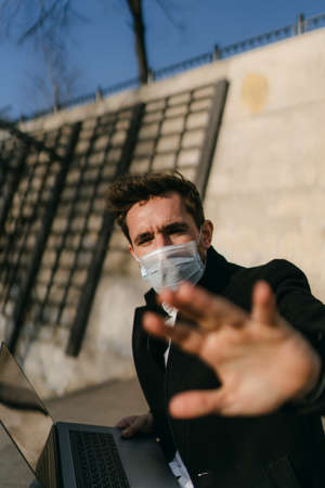 A man sits on the street wearing a medical mask and works behind a laptop during coronavirus quarantine and emotionally asks not to shoot himselfの写真素材