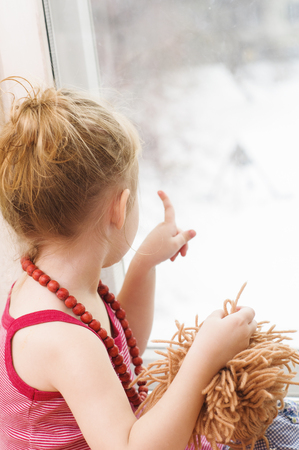 Little girl with doll looking out the window of the houseの写真素材