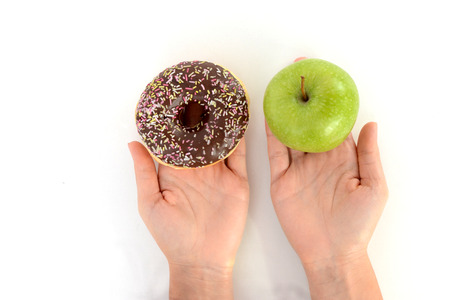 Dieting concept with young woman choosing between healthy fruits and sweets, holding green apple and tasty chocolate donutの写真素材