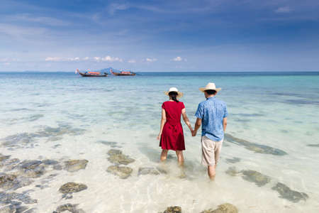Couple holding hand walking in to crystal clear sea with long tail boat background, Krabi, Thailandの写真素材