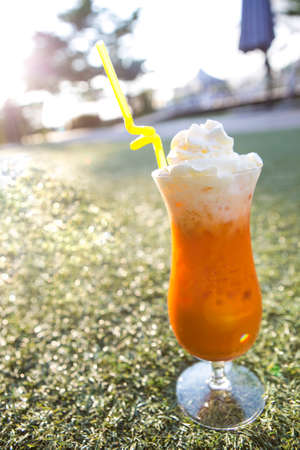 orange thai tea with ice top with whipped cream serve in tall glass and straw on a green grassの写真素材