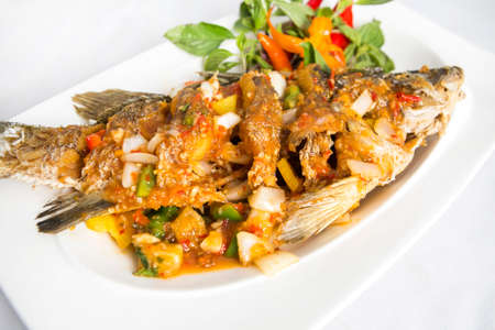 fired sea bass top with thai spicy sauce decorated with basil leaves and chilis served in white plateの写真素材