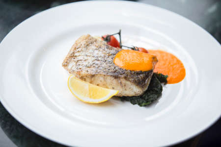 grilled seabass dish served with orange sauce, tomato, spinach and a piece of lemon put on white...