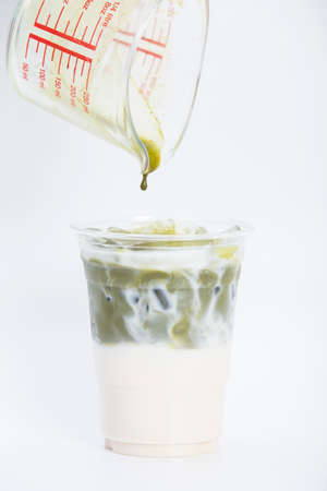 a drop of intense mixed matcha green tea latte, from measuring beaker glass on topped of fresh milk in takeaway plastic cup isolated white backgroundの写真素材