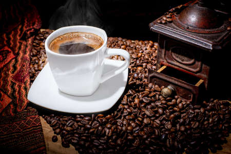 White cup of steaming coffee. A wooden table covered with roasted seeds.. Vintage, retro bronze coffee mill on black background. Brown coffee beans. Cashmere element. Rustic style.の写真素材
