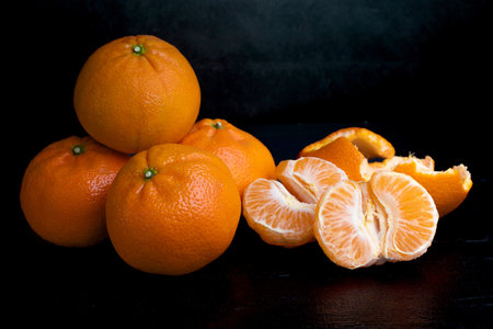 A stack of four tangerines on a dark background. Orange fruits on a black background. Citrus in the kitchen. A stack of four tangerines on a dark background. Juicy fruits. One peeled tangerineの写真素材