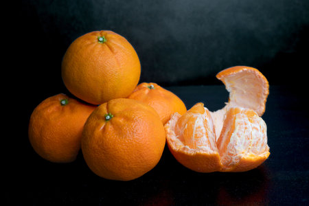 A stack of four tangerines on a dark background. Orange fruits on a black background. Citrus in the kitchen. A stack of four tangerines on a dark background. Juicy fruits. One peeled tangerineの写真素材