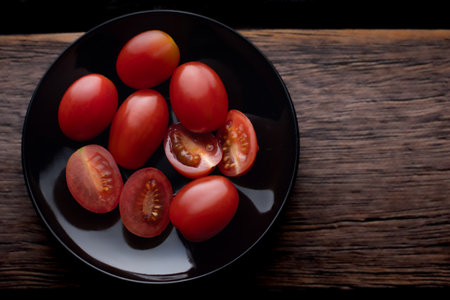 Cherry tomatoes on a black round plate. Dark background. Brown boards. Spices in the kitchen.の写真素材