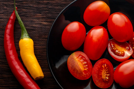 Cherry tomatoes, red and yellow peppers on a black round plate. Dark background. Brown boards. Spices in the kitchen.の写真素材
