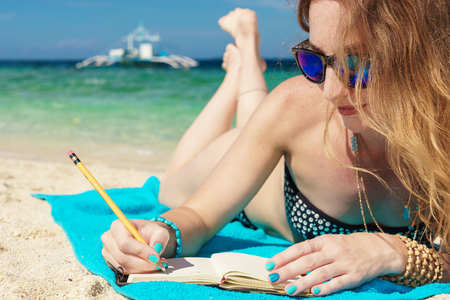 young european woman with sunglasses is lying on the coast of tropical turquoise sea and wrigting by pencil in notepad at sunny summer day under blue sky.の写真素材