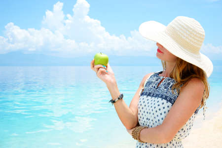 Young beautiful european woman is holding green apple in her hand against the calm soft tropical sea under tender blue sky at sunny dayの写真素材