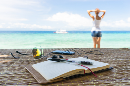 Relaxing girl staying on the beach. Opened empty notepad is on the table with sunglasses, phone and headphones at the tropical sea background and philippine boat on his surface. At sunny day under cloudy skyの写真素材