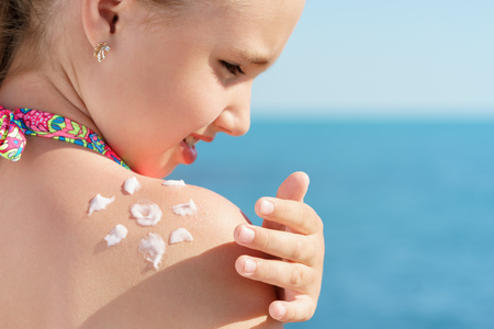 young girl applyng sun protector cream on her shoulder on the beach close to tropical turquoise sea under blue sky at sunny dayの写真素材
