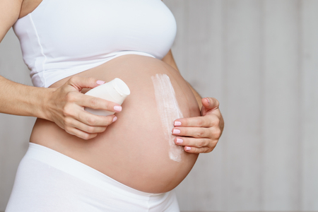 Pregnant woman aplying cream at her belly for prevention of stretch marks on the skin in light roomの写真素材
