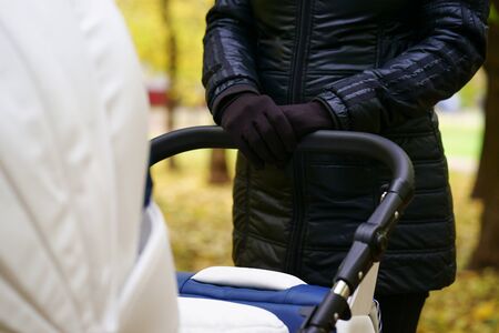 young caucasian woman is pushing white baby carriage while she walking at autumn park with beautiful yellow leaves at day timeの写真素材