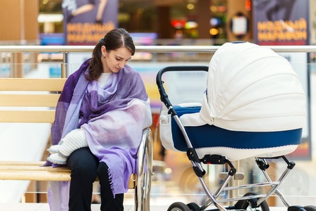 Young european woman with violet stole is breastfeeding her little child close to white baby carriage at public place shopping mall at day timeの写真素材