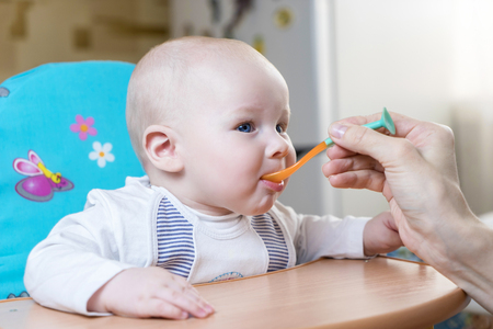 Mom feeds by spoon an emotional infant boy with blue eyes in kitchen at day timeの写真素材