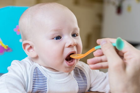 Mom feeds by spoon an emotional infant boy with blue eyes in kitchen at day timeの写真素材