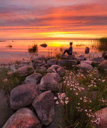 Fiery sunset with the girl, flowers and stones on the lakeの写真素材