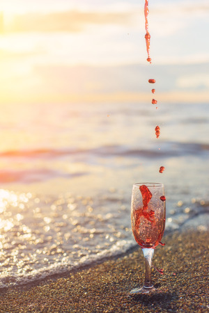 A tall glass with splashes of red wine on the seashore. Sunsetの写真素材